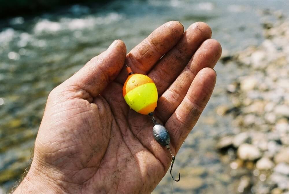 Zbliżenie na dłoń wędkarza prezentującą pękaty spławik rzeczny i obciążenie na tle wody.
