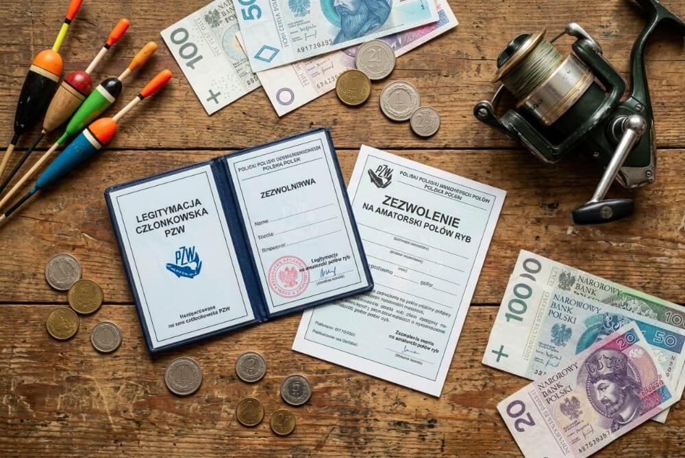 Dokumenty wędkarskie oraz polskie pieniądze leżące na stole obok sprzętu wędkarskiego.