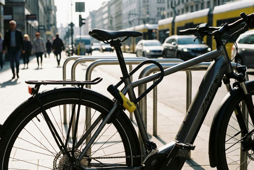 Rower trekkingowy przypięty solidnym zapięciem typu U-lock do miejskiego stojaka na tle ulicy.